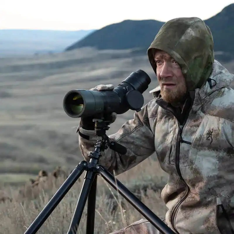 A man in camouflage outerwear using a spotting scope on a tripod.